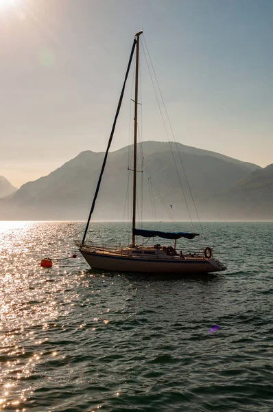 Sailing yacht floating near shore of misty Garda lake with high dolomite mountains with sun shining above in the sky on the background. Northern Italy, Lombardy — Stock Photo, Image