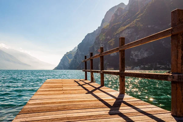 Naturskön utsikt över träplankor piren med räcken byggda på norra stranden av vackra Gardasjön i Lombardiet, Italien omgiven av höga dolomitberg och kristallklart blått vatten i sjön. Riva Stockfoto