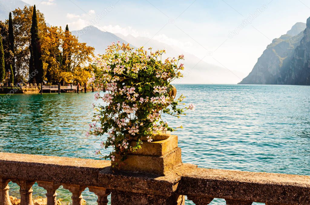 Beautiful Garda lake promenade with classic stone fence railings built on the edge with flowerpots with blooming white flowers. Garda lake surrounded by the high dolomite mountains on the background