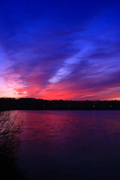 湖の上にカラフルな夕日 水に反射するピンクと青の積乱雲 前景に植生のシルエット 背景の街の光 — ストック写真