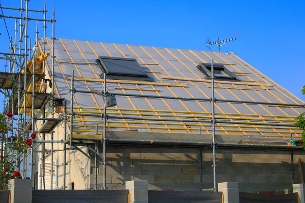 Roof frame under construction. Tile bedding with steam and rain barrier protection. Roof window with drop-down blind. Scaffolding against the walls of the front of the house.