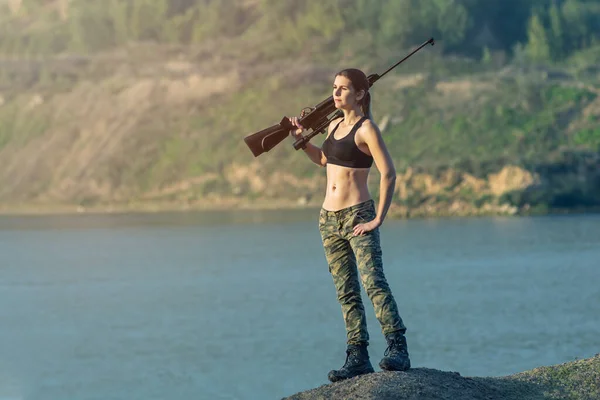 Menina de moda militar com rifle com uma visão óptica. Feminino caçador de esportes de perseguição na floresta com arma . — Fotografia de Stock