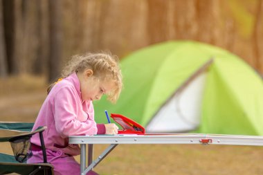 Yakın plan küçük kız açık havada çadırın yanındaki bir masada bir sandalyeye oturur ve ders verir. Çocuklar için uzaklık öğrenme kavramı