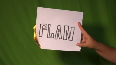 Bir el, arkasında yeşil ekran üzerinde plan metni olan yanan kağıdı tutar..