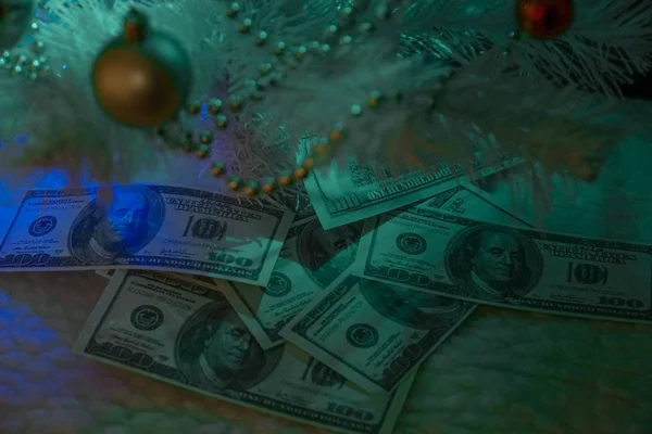 Dolarlar yeni yıl sepetinin altında. Noel ağacının altında bir hediye. Noel süslemeli beyaz Noel ağacı. Dolarlarla iş. Masaya yığınla para..