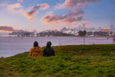 Crissy Field 'dan San Francisco' da gün batımını seyreden orta boy bir çift fotoğrafı.