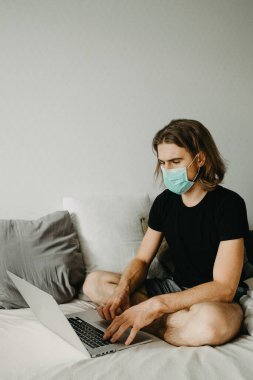 Evde karantina sırasında dizüstü bilgisayar kullanan beyaz bir hippi. Beyaz adam evde uzaktan çalışıyor ve virüs önleme için maske takıyor Coronavirus konsepti.. 
