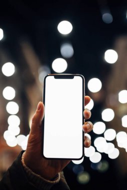 Kafkasyalı bir kadının elindeki boş beyaz cep telefonu ekranına yakın çekim. Parlak, arka planda Bokeh City ışıkları bulanıklaştı. Mobil uygulama şablonu.