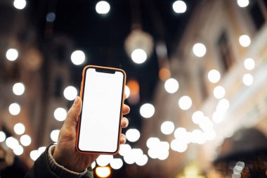 Kafkasyalı bir kadının elindeki boş beyaz cep telefonu ekranına yakın çekim. Parlak, arka planda Bokeh City ışıkları bulanıklaştı. Mobil uygulama şablonu.