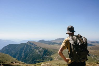 Sırt çantalı genç hippi güneşli bir günde dağ manzarasının tadını çıkarıyor. Dağda yaşayan bir yürüyüşçü. Boş bulutsuz gökyüzü.