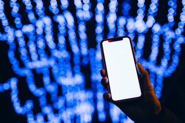 Kafkasyalı bir kadının elindeki boş beyaz cep telefonu ekranına yakın çekim. Arka planda Bokeh şehrinin ışıklarını bulanıklaştıran mavi ışıklar var. Mobil uygulama şablonu.
