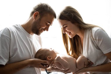 Beyaz tişörtler giyen, küçük kızlarını kucağında tutan, ona bakıp gülümseyen mutlu bir aile. Arka planda duygusal aile anı.