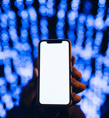 Kafkasyalı bir kadının elindeki boş beyaz cep telefonu ekranına yakın çekim. Arka planda Bokeh şehrinin ışıklarını bulanıklaştıran mavi ışıklar var. Mobil uygulama şablonu.