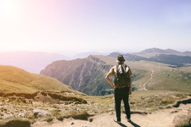 Sırt çantalı genç hippi güneşli bir günde dağ manzarasının tadını çıkarıyor. Dağda yaşayan bir yürüyüşçü. Boş bulutsuz gökyüzü.