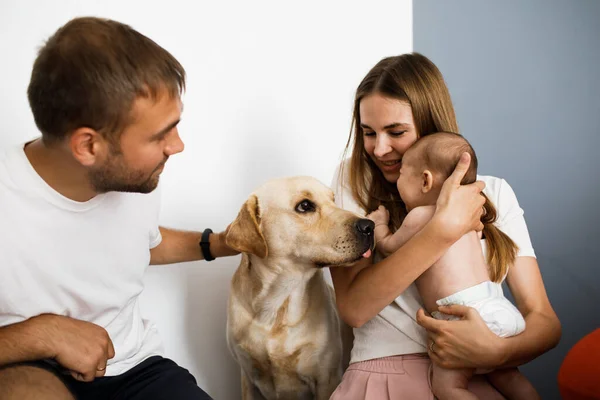 Happy family with little baby and big dog spend time together. Mother holding and hugging her daughter. Parents smiling and looking at their pet. Staying home.