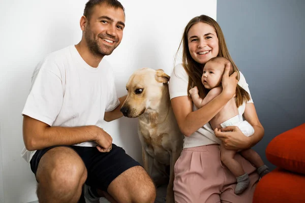 Happy family with little baby and big dog spend time together at home. Parents similing and looking straight into the camera. Staying at home.