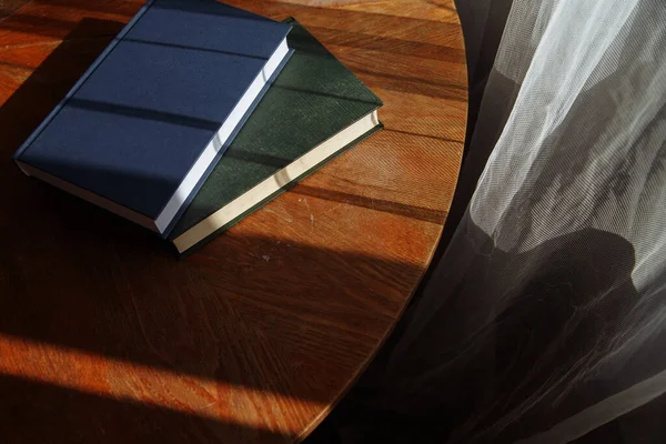 two books green and blue on a wooden table and a shadow from the curtains