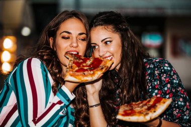 İki genç kadın dışarıda pizza yiyor, birlikte eğleniyorlar.