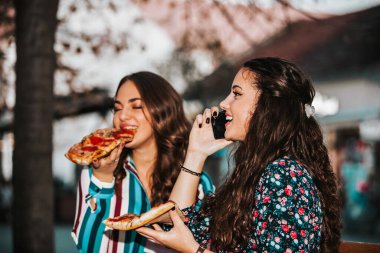 Two beautiful female friends talking and eating pizza outdoors.