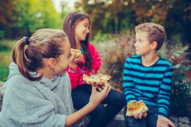 Three happy children talking and eating pizza outdoors, having f