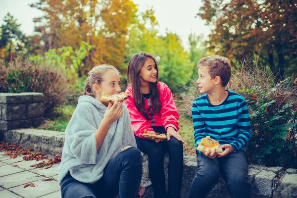 Three happy kids talking and eating pizza outdoors — Stok fotoğraf