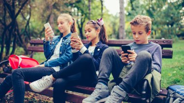 Group of friends hang out after school and using smart phone while sitting on the bench in the park.