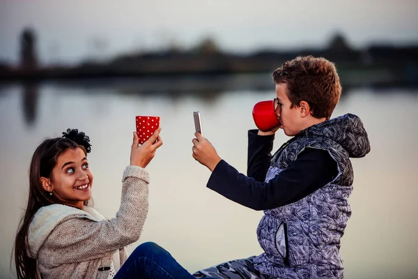 夕方に湖のそばに座っている間 楽しさとお茶を飲んでいる男の子と女の子の2人の若いかわいい友達 男の子はお茶のカップの彼の携帯電話で写真を撮る — ストック写真