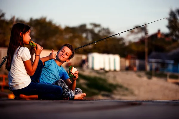 两个年轻可爱的小朋友 男孩和女孩在阳光明媚的夏日吃三明治 在湖里钓鱼 男孩把手势大拇指推上去 孩子们在玩友谊 — 图库照片