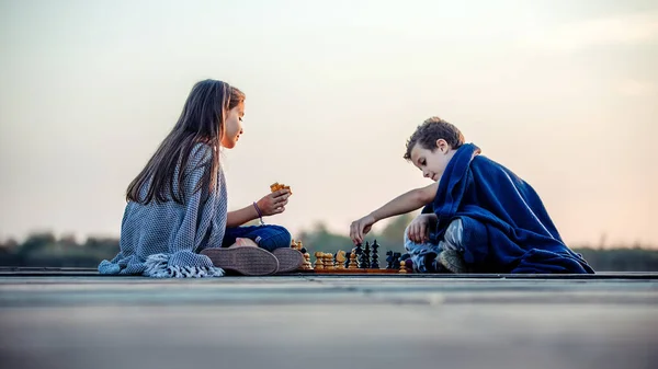 Two young happy little friends, boy and girl having fun while playing chess sitting by the lake in the evening. Kids are playing. Friendship.