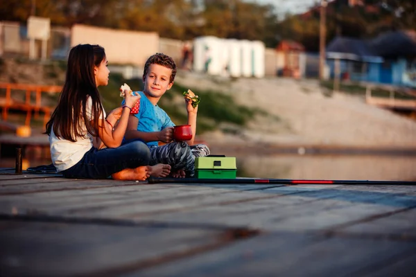 Two young cute little friends, boy and girl talking, drinking tea, eating sandwiches and fishing on a lake in a sunny summer day. Kids are playing. Friendship.