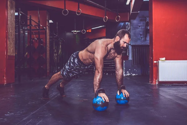 Young muscular bearded man with naked torso doing push ups with kettlebell weights in the gym