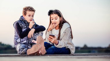 İki küçük sevimli arkadaş, erkek ve kız gece gölün kenarında oturmuş akıllı telefondan bir şey izliyorlar. Çocuklar birlikte eğleniyor. Arkadaşlık.