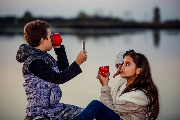 夕方に湖のそばに座っている間 楽しさとお茶を飲んでいる男の子と女の子の2人の若いかわいい友達 女の子はお茶のカップの彼女の携帯電話で写真を撮る — ストック写真