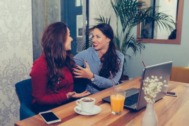 İki kadın arkadaş dizüstü bilgisayar kullanıyor ve birlikte kahve içiyor. Kafede konuşan, gülen ve eğlenen iki kadın.
