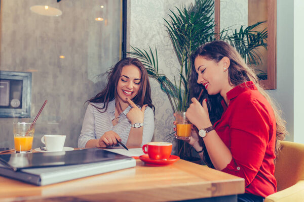 Happy female friends, students reading notes together, pointing where to sign a contract, talking and drinking coffee while sitting in the table in cafe. - Image