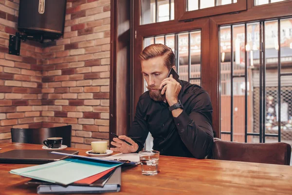 Successful bearded business man writing document during mobile phone conversation with client, sitting at table in cafe. Male skilled financier having cell phone conversation.