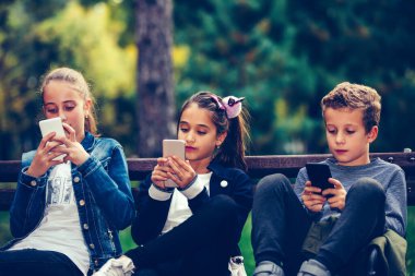 Group of friends playing video games on smart phone after school while sitting on the bench in the park.