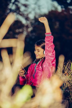 Excited little girl dancing, singing and listening music with headphones and smart phone in the park