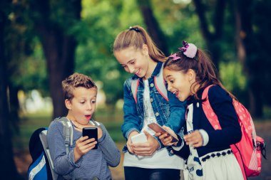 Bir grup gülen arkadaş okuldan sonra takılır ve parkta yürürken akıllı telefon kullanır. Çocuklar gülüyor ve telefonla eğleniyorlar. Seçici odak