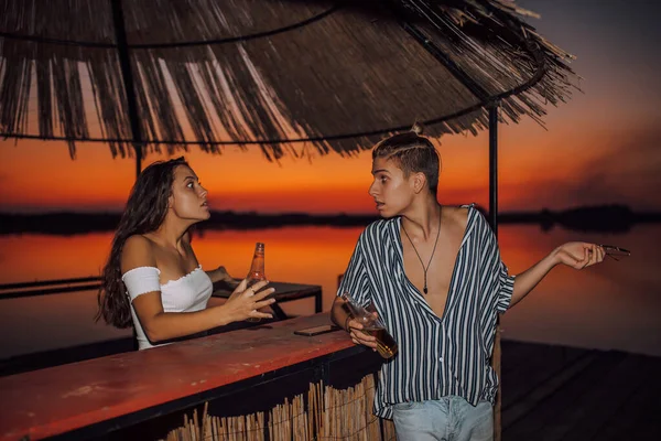 若いカップルはドリンクと議論とビーチバーで時間を過ごしています 関係の問題だ 男は動揺した女の子に何かを説明する — ストック写真