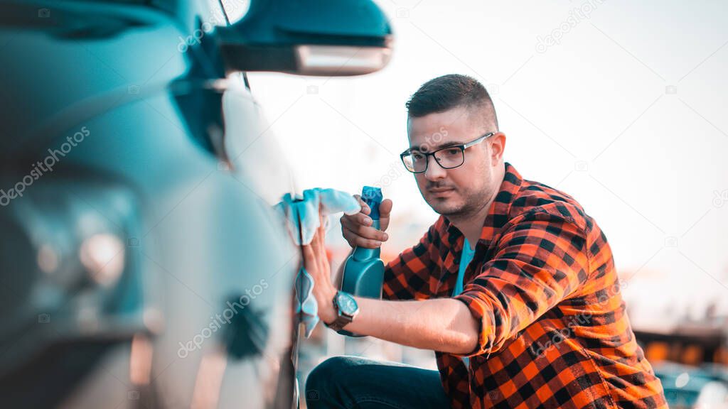 Portrait of a young man cleaning his car with a cloth and spray in a bottle, outdoors.