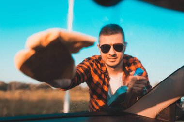 Genç adam arabasının ön camını mikrofiber bezle dikkatlice temizliyor ve şişeye sıkıyor..