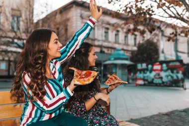 Dışarıda pizza yiyen ve arkadaşlarına el sallayan iki güzel genç kadın. Yaşam tarzı, modern hayat..