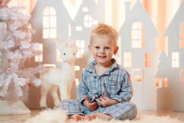 Sevimli sarı saçlı küçük çocuk pijama yakınındaki Noel oyuncak kağıt evler.