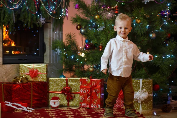 Cute blonde hair little boy near the fireplace and gifts under Christmas tree.