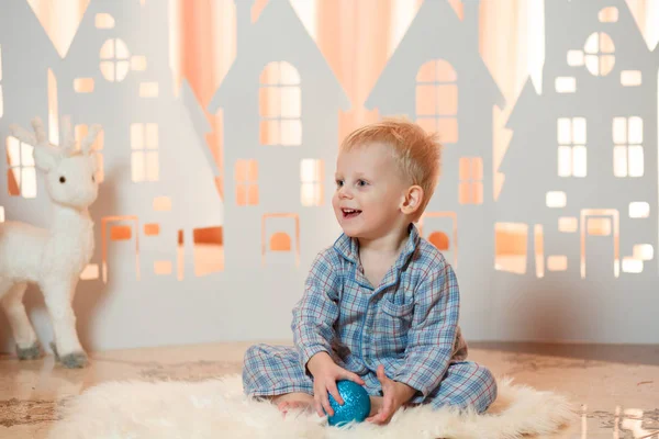 Cute blonde hair little boy in sleepwear near christmas toy paper houses.