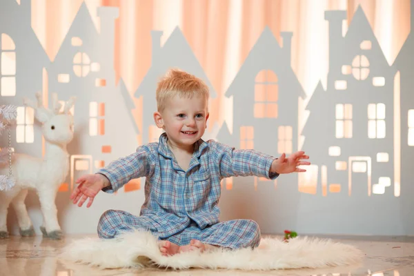 Cute blonde hair little boy in sleepwear near christmas toy paper houses.