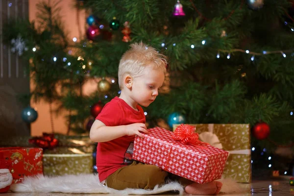 Cute blonde hair little boy near the fireplace and gifts under Christmas tree.