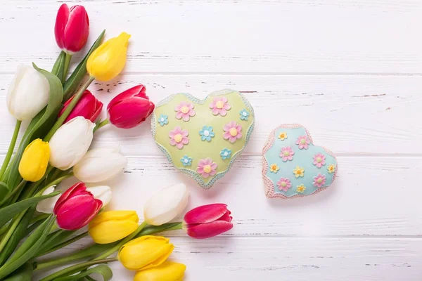 Yellow, red and white tulips flowers and two hearts on white wooden background