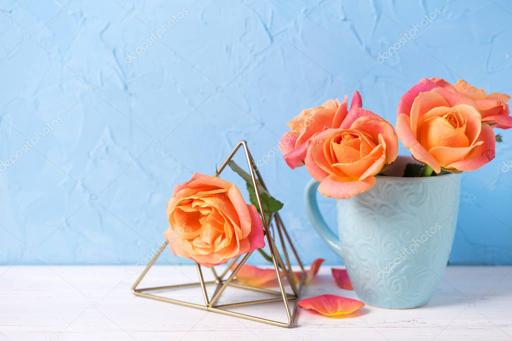 Floral still life. Bunch of fresh orange roses  in cup  on white wooden background against  blue wall. Place for text. 